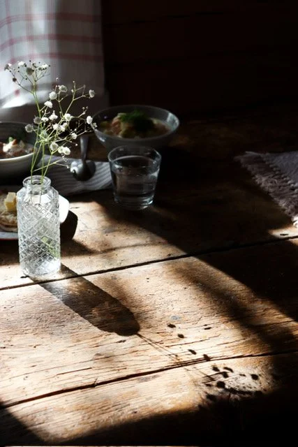 A small glass vase with white flowers, a glass of water, and a bowl of food on a rustic wooden table with sunlight casting shadows.
