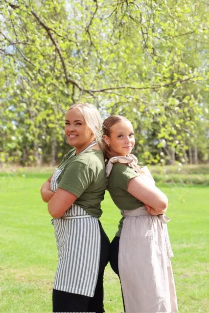 Two women standing back to back outdoors in a grassy area with trees and green leaves in the background, smiling at the camera.