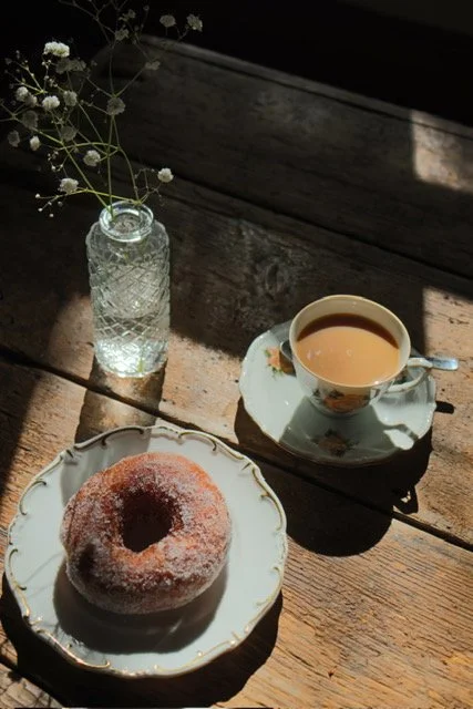 A plate with a sugar-coated donut, a cup of tea or coffee with milk on a saucer, a small glass bottle with white baby's breath flowers, all placed on a rustic wooden table.