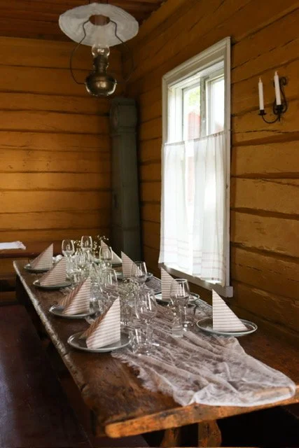 Dining table set with plates, glasses, and napkins in a rustic wooden room with a window and wall-mounted candle holder.