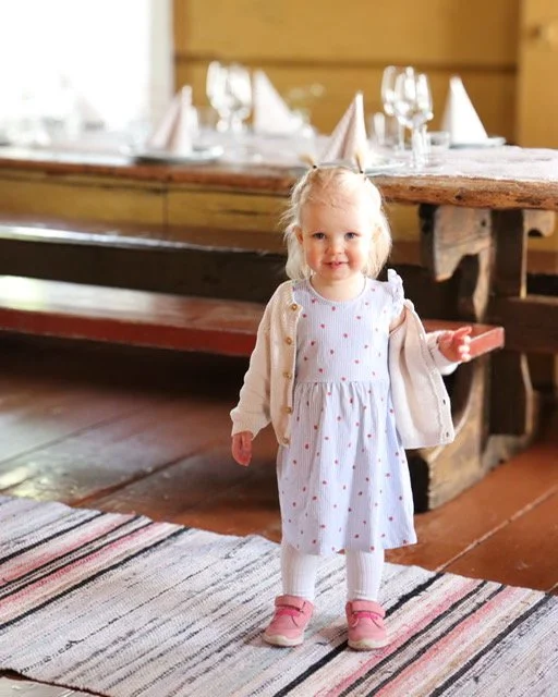 A young girl with blonde hair and a light-colored dress with small red dots, standing on a rug in a rustic dining area, holding one arm out, with a bubblegum pink shoe on her right foot. In the background, there is a wooden dining table set with wine glasses and folded napkins.