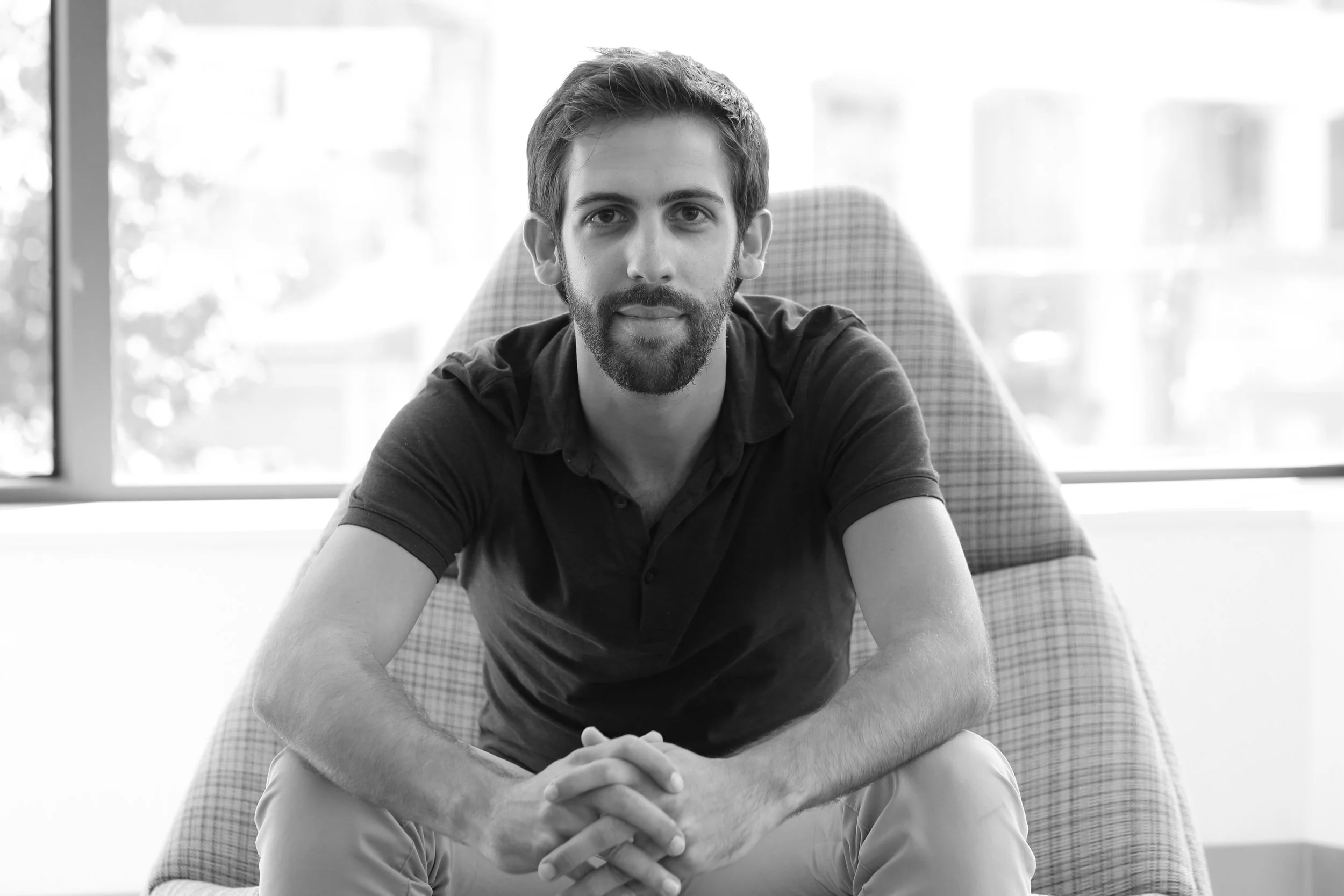 A man with a beard and short hair sitting on a checkered armchair in front of a window, looking at the camera.