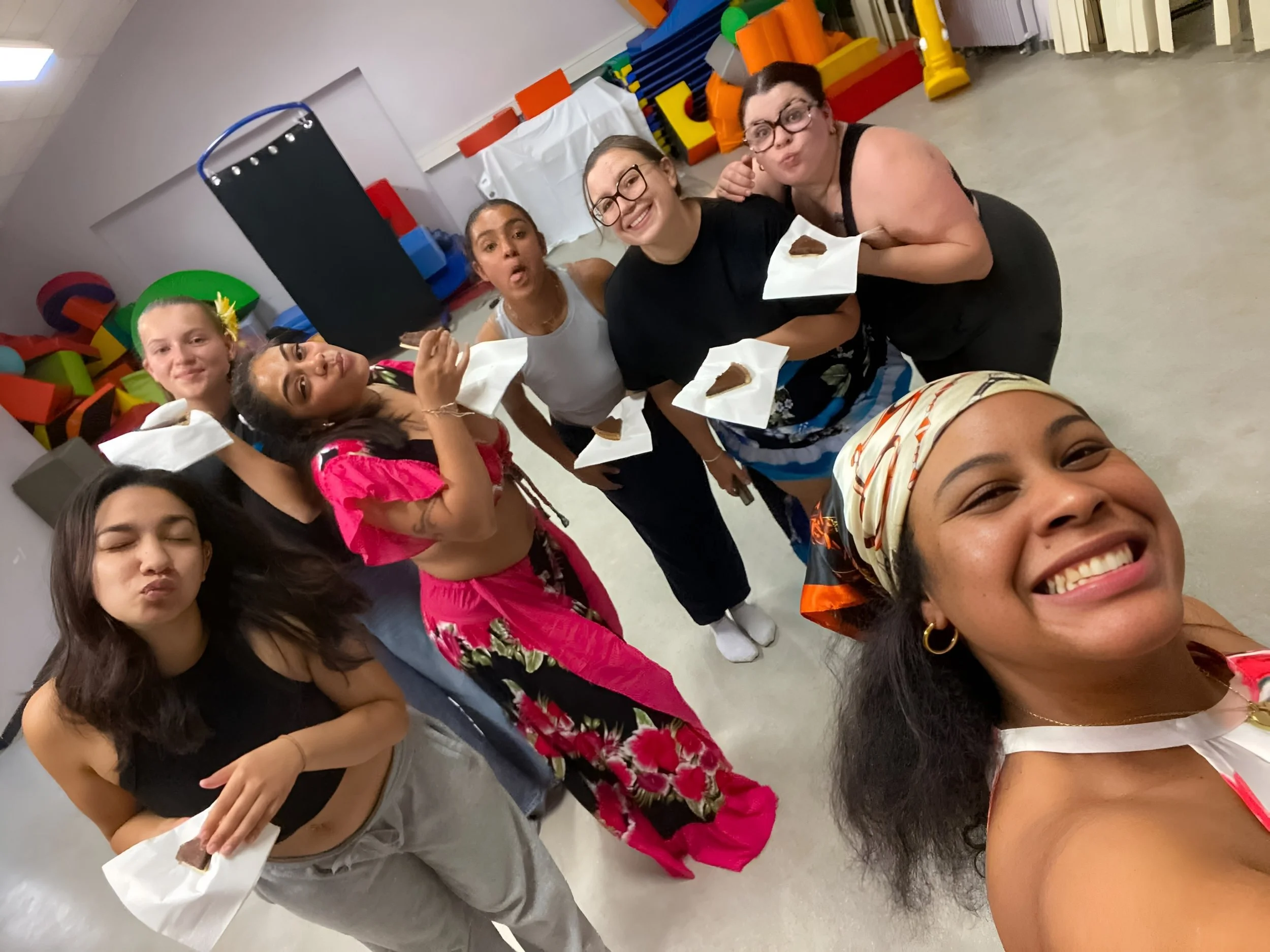 Groupe de cinq femmes souriantes et détendues, tenant des morceaux de gâteau emballés dans du papier blanc, dans une salle colorée de jeux et d'activités pour enfants.