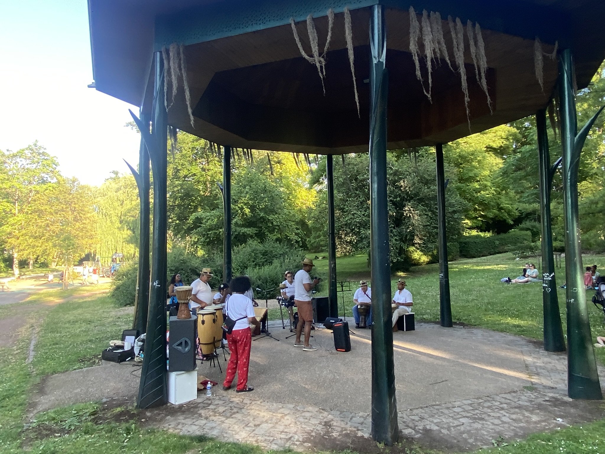 Groupe de personnes jouant de la musique dans un parc lors d'un événement en plein air, sous une structure en bois avec piliers métalliques, entourée d'arbres verts en été.