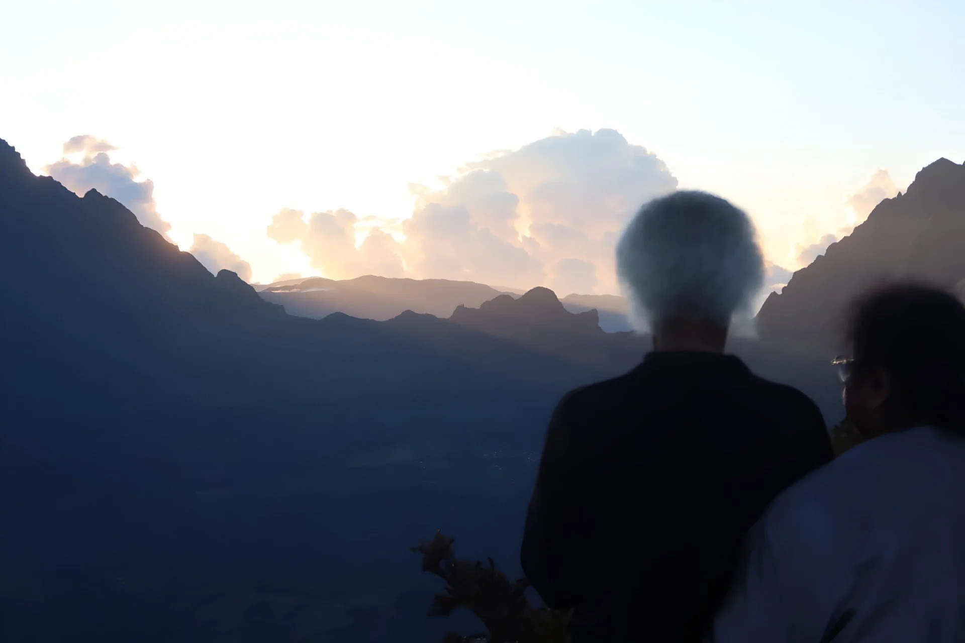 Deux personnes regardant un paysage de montagnes au coucher du soleil, avec des nuages dans le ciel.
