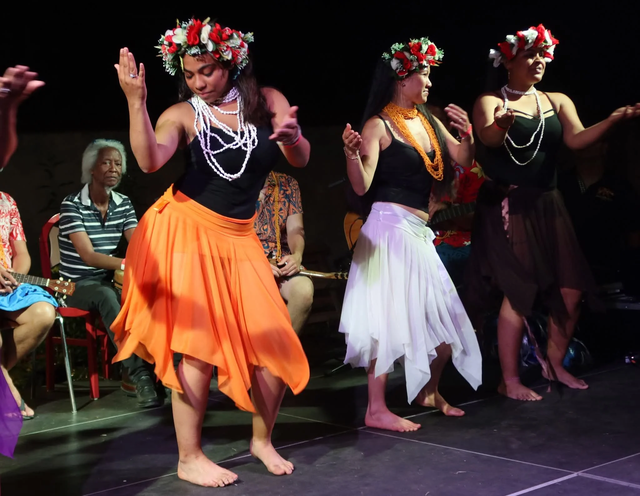 Femme dansant traditionnellement avec un bandeau de fleurs sur la tête, vêtue d'une jupe orange et d'un haut noir, accompagnée d'autres femmes dans des tenues similaires et un homme assis en arrière avec une guitare. La scène semble être un spectacle culturel ou une performance artistique.
