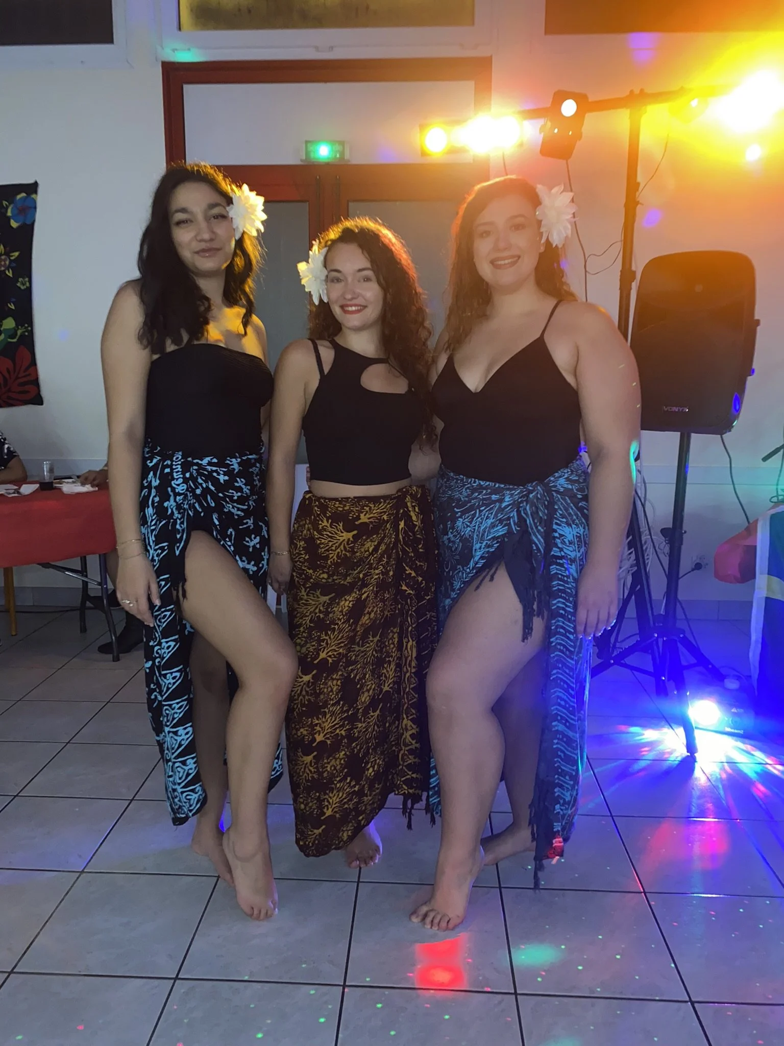 Trois femmes en maillot de bain avec des fleurs dans les cheveux posant pour la photo lors d'une fête avec un éclairage coloré et un système de son sur un mur blanc.