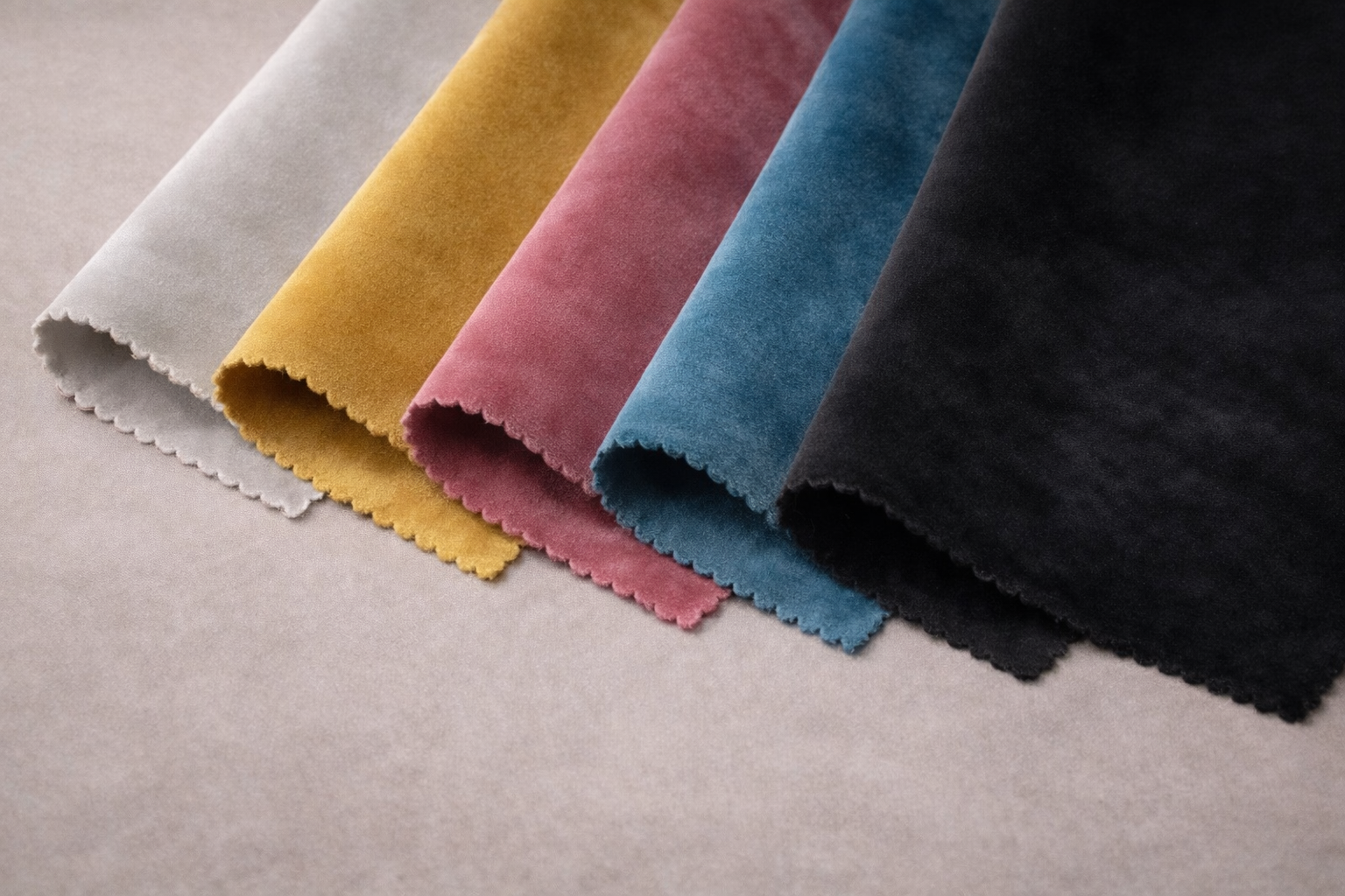Muestra cinco telas de diferentes colores: gris claro, mostaza, rosa, azul y negro, dobladas y colocadas una al lado de la otra en una superficie de color gris.