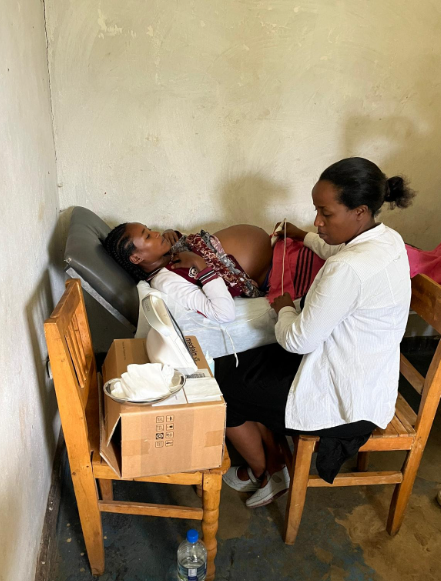 En kvinna som är gravid lying på en sjukhussäng, medan en annan kvinna står bredvid och tar hand om henne. Rummet är enkelt och har trästolar, ett vattenflaska på golvet, och medicinska och förvaringsartiklar på ett bord.
