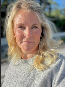En kvinna med blont hår och blå ögon utomhus, solen lyser på hennes ansikte.