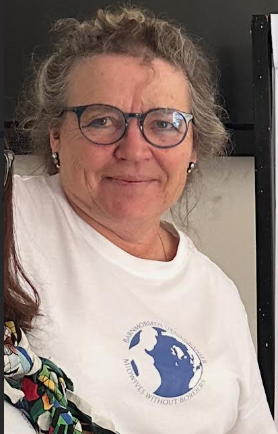 En kvinna med lockigt grått hår, glasögon och vit T-shirt, står inomhus.