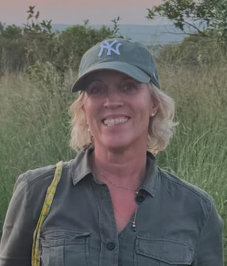 En kvinna utomhus, bär en grå New York Yankees-keps och en mörkjeansjacka. Hon ler mot kameran, omgiven av grön vegetation.