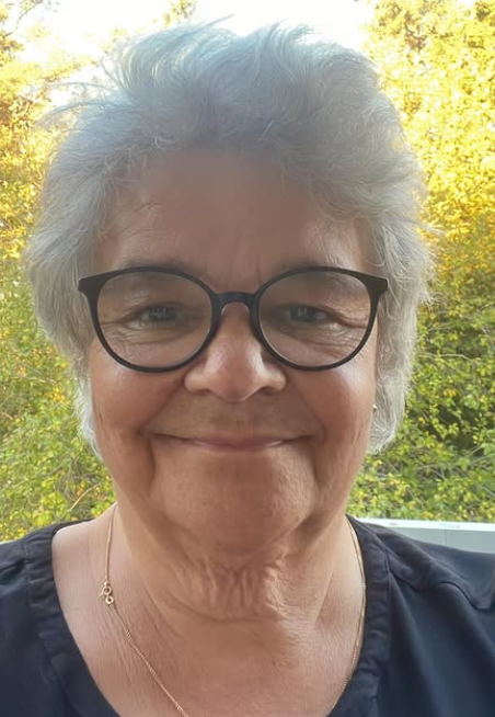 En äldre kvinna med kort, grått hår och svarta glasögon, utomhus med gröna träd i bakgrunden, ler och tar selfie.