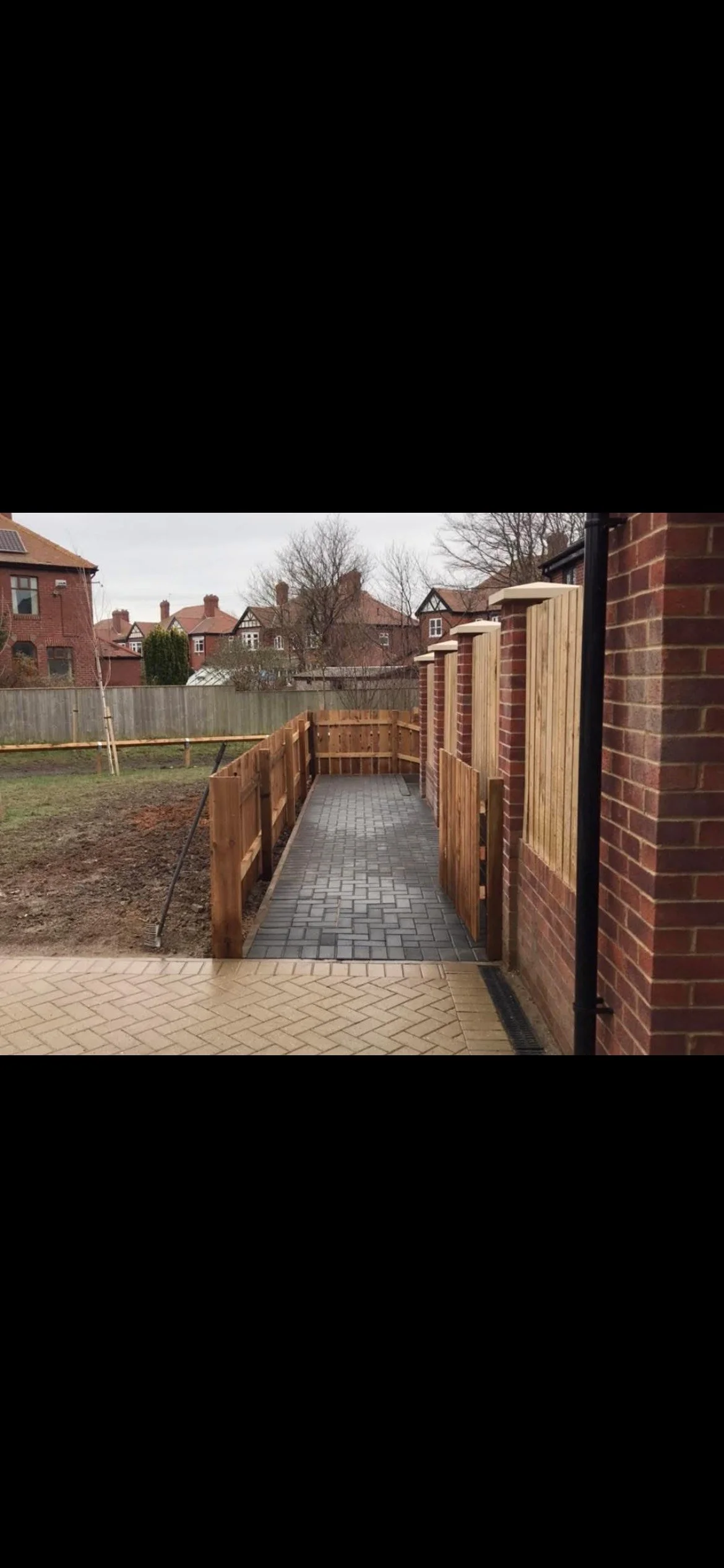 Newly constructed wooden fenced pathway extending from a brick house to a backyard with neighboring houses in the background.