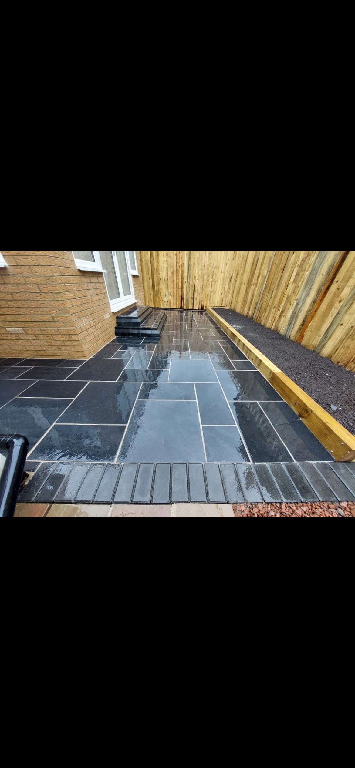 Backyard patio with black polished tiles, wooden steps, brown brick house, and a new wooden fence with a narrow planter box.