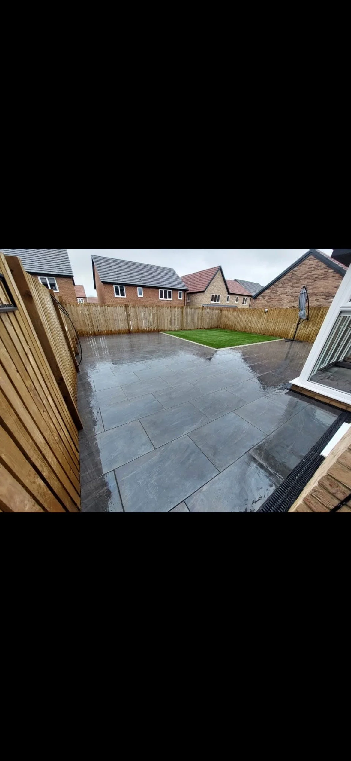 Backyard with wet gray tiled patio, small patch of green grass, wooden fence, and neighboring houses in the background.