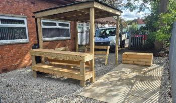 New wooden outdoor seating area with a bench, table, and planter on gravel and concrete ground near a brick building with windows.