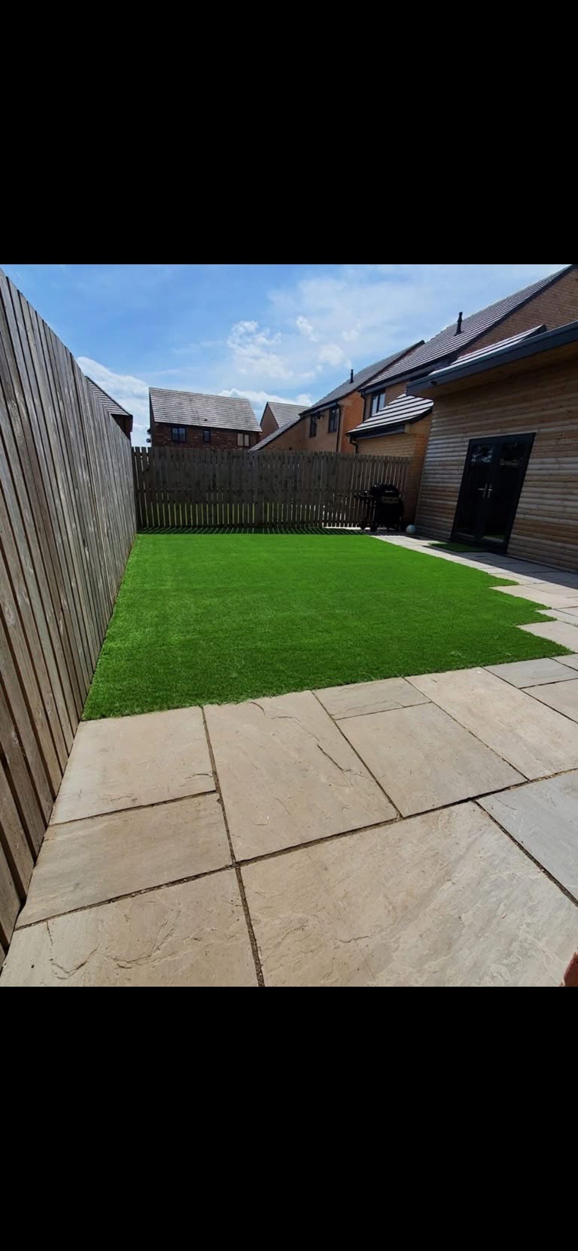 A backyard with a stone patio area, a strip of grass, wooden fencing on two sides, and neighboring houses in the background under a partly cloudy sky.