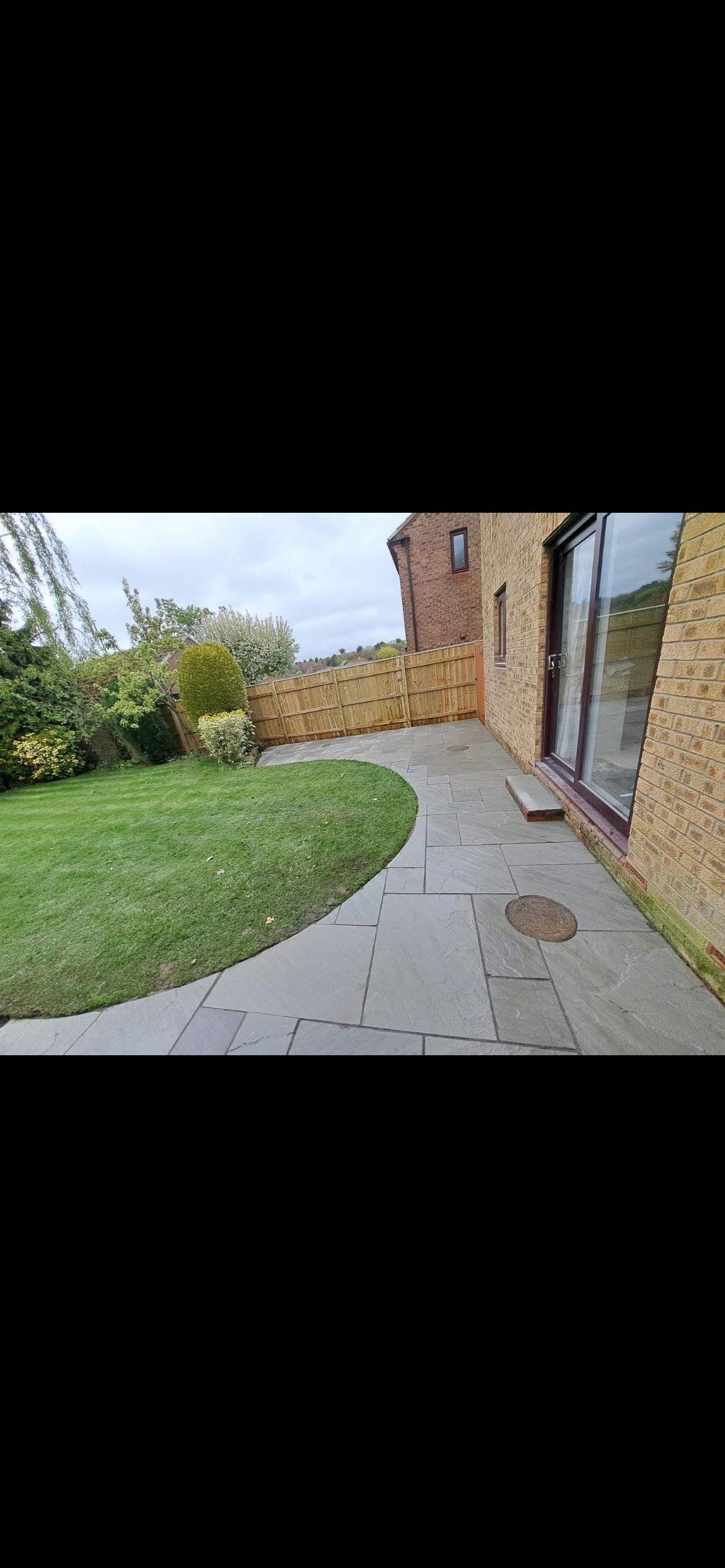 Backyard patio with gray stone paving, green grass, a wooden fence, bushes, and trees, adjacent to a brick house with sliding glass doors.