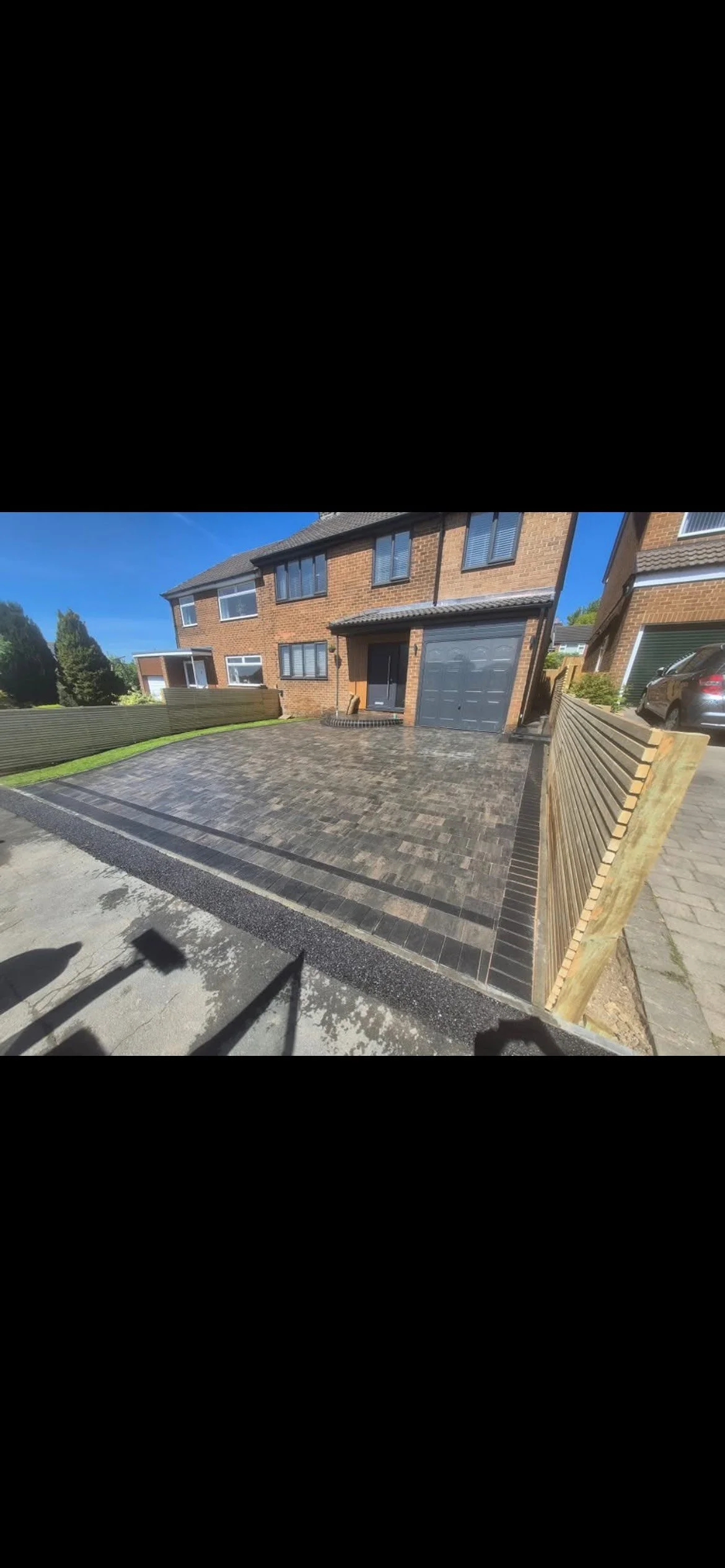 Newly paved driveway with gray and black interlocking bricks in front of a brick house with black garage door, surrounded by wooden fencing and neighboring houses.