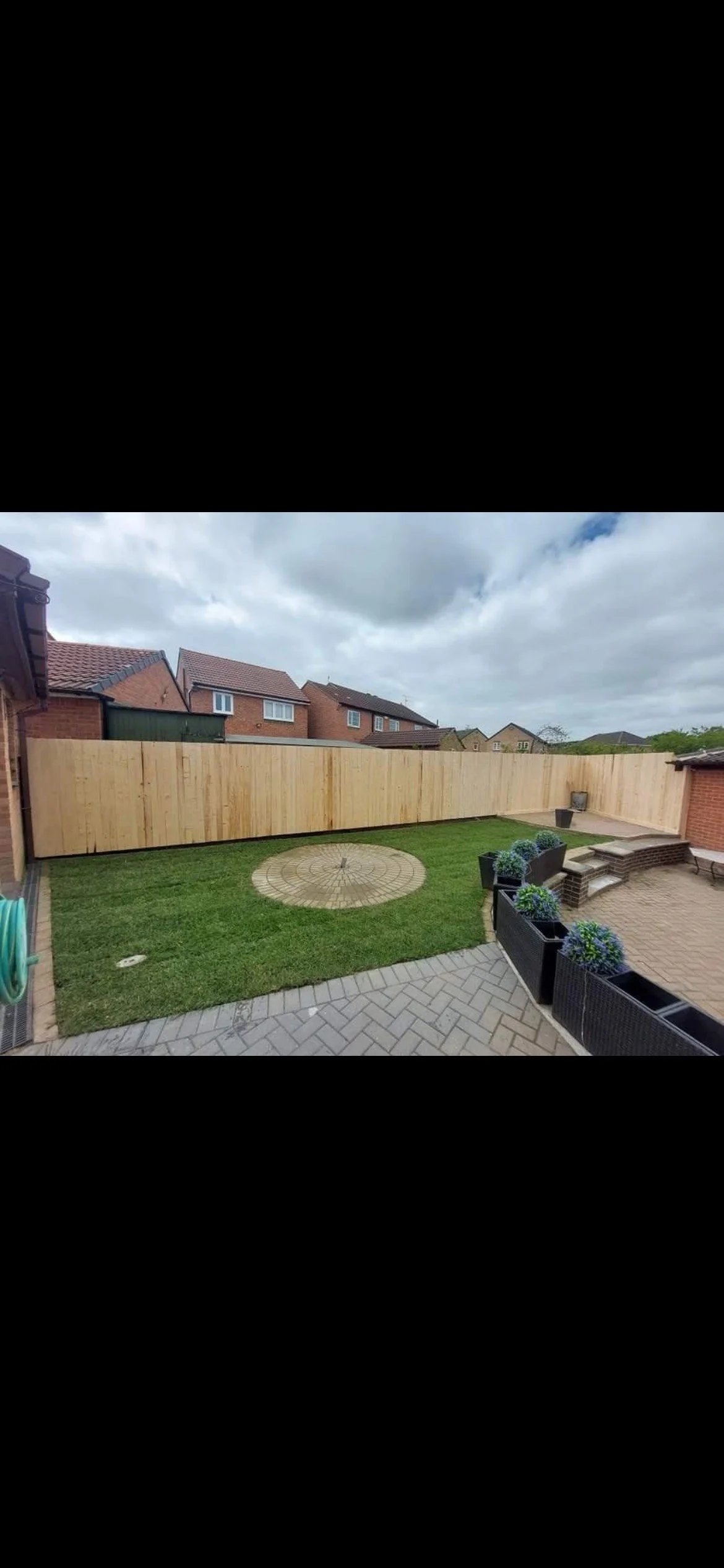 Backyard with a wooden privacy fence, a grassy area with a circular brick patio, potted plants along the patio, and neighboring houses with cloudy sky overhead.