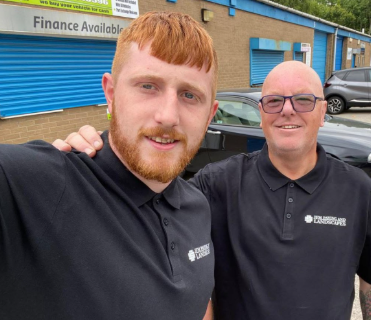 Two men taking a selfie outdoors, both wearing black polo shirts with a logo, standing in front of a blue building with a sign that reads 'Finance Available'.