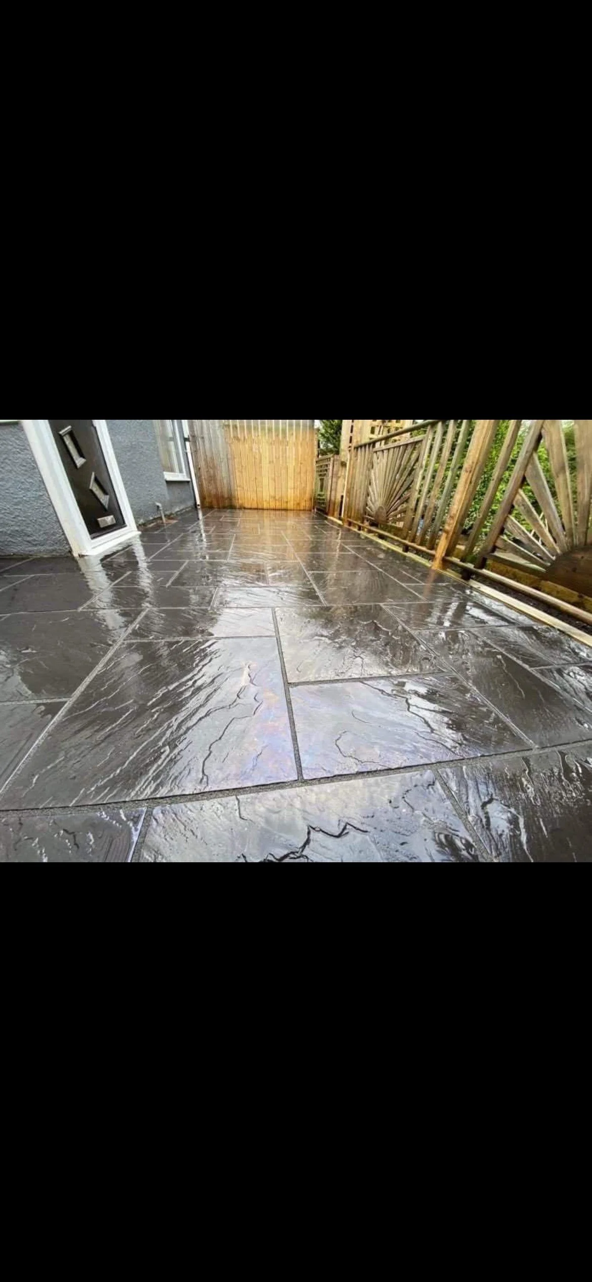Photo of a wet outdoor patio with dark stone tiles, bordered by a wooden fence on the right and a gray house wall with a white door on the left.