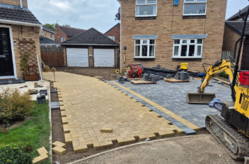 A construction site showing a partially completed brick pathway in a backyard, with construction equipment and building materials around.