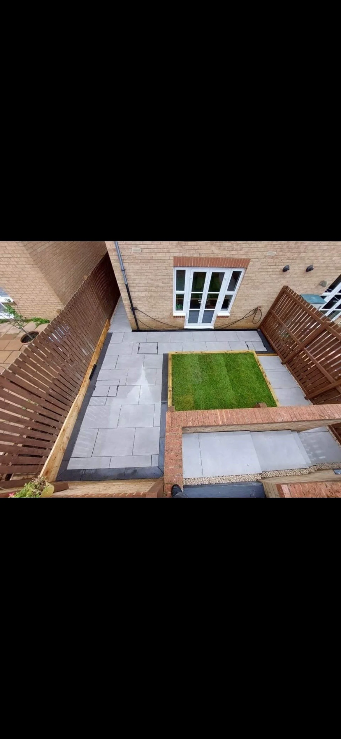 A small backyard patio with a green grass patch, paved stone pathway, and wooden fencing, connected to a brick house with a glass door.