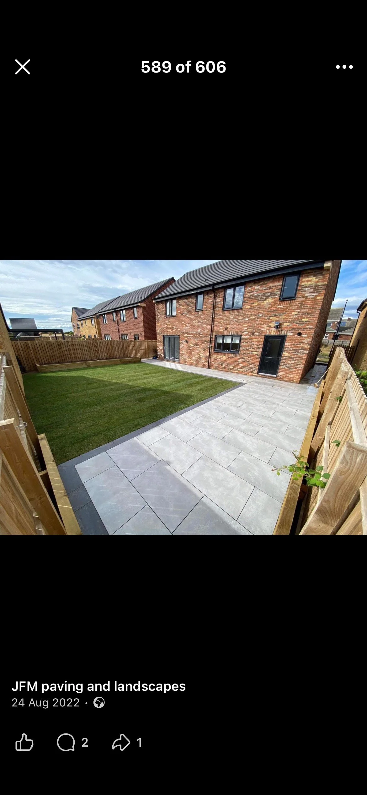 Backyard with a grassy lawn area enclosed by a wooden fence, adjacent to a patio with large gray tiles, and a brick house with multiple windows and a black door under a partly cloudy sky.
