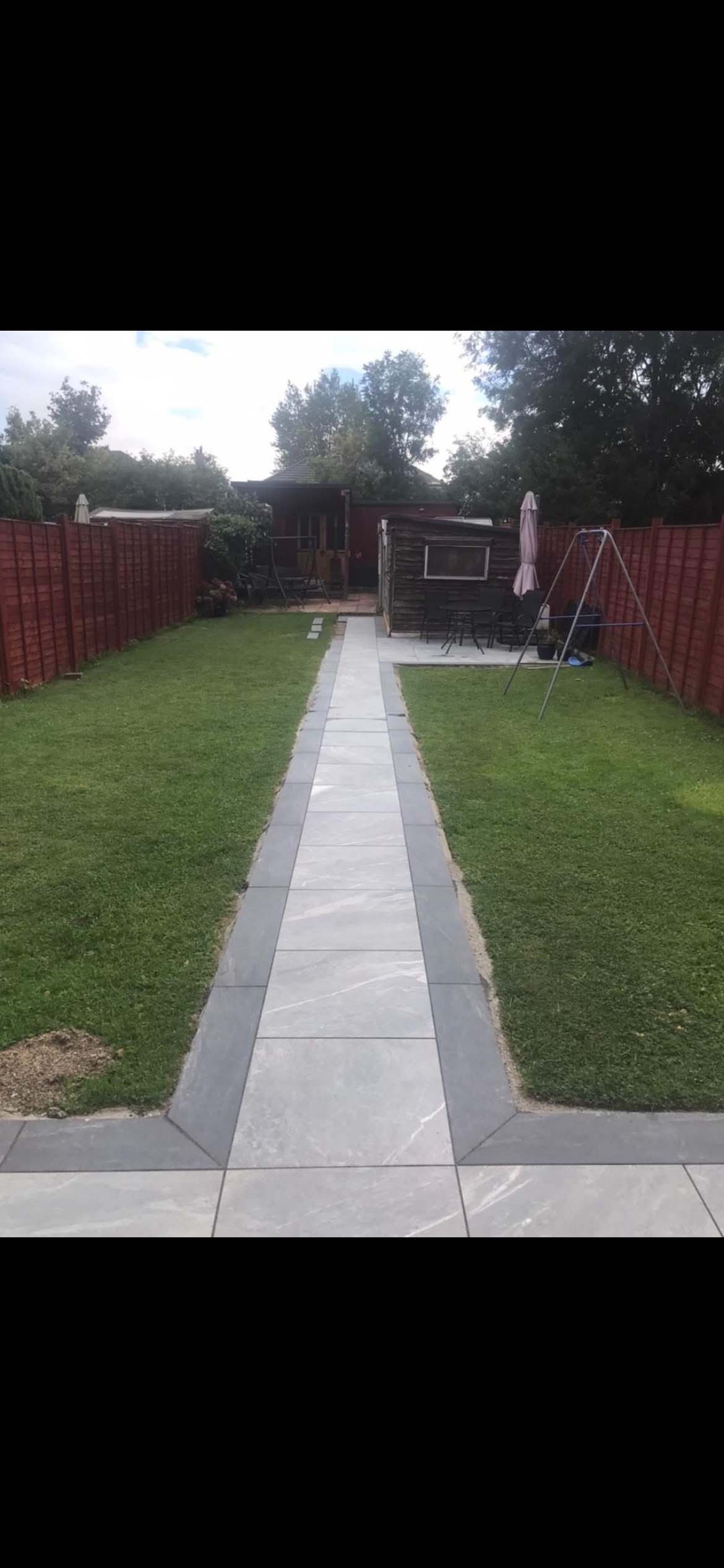 View of a backyard with a paved walkway, green grass on either side, red wooden fence, garden shed, outdoor dining area, and swing set.