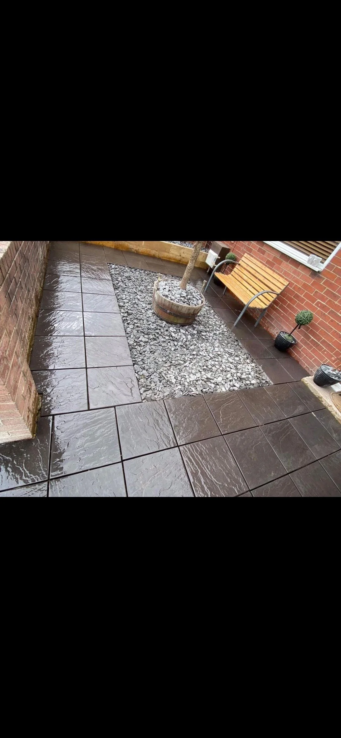 An outdoor patio area with wet dark brown tiled flooring, a small tree in a wooden barrel planter, a wooden bench, and potted plants against a brick wall.