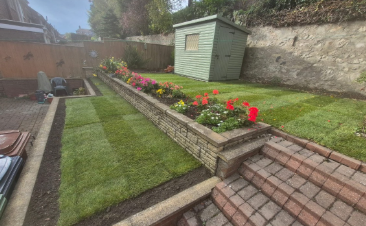 A backyard garden with neatly trimmed grass, colorful flowers along a brick border, and a small green shed in the background. There's a stone path leading to the shed and a red-tiled roof in the foreground.