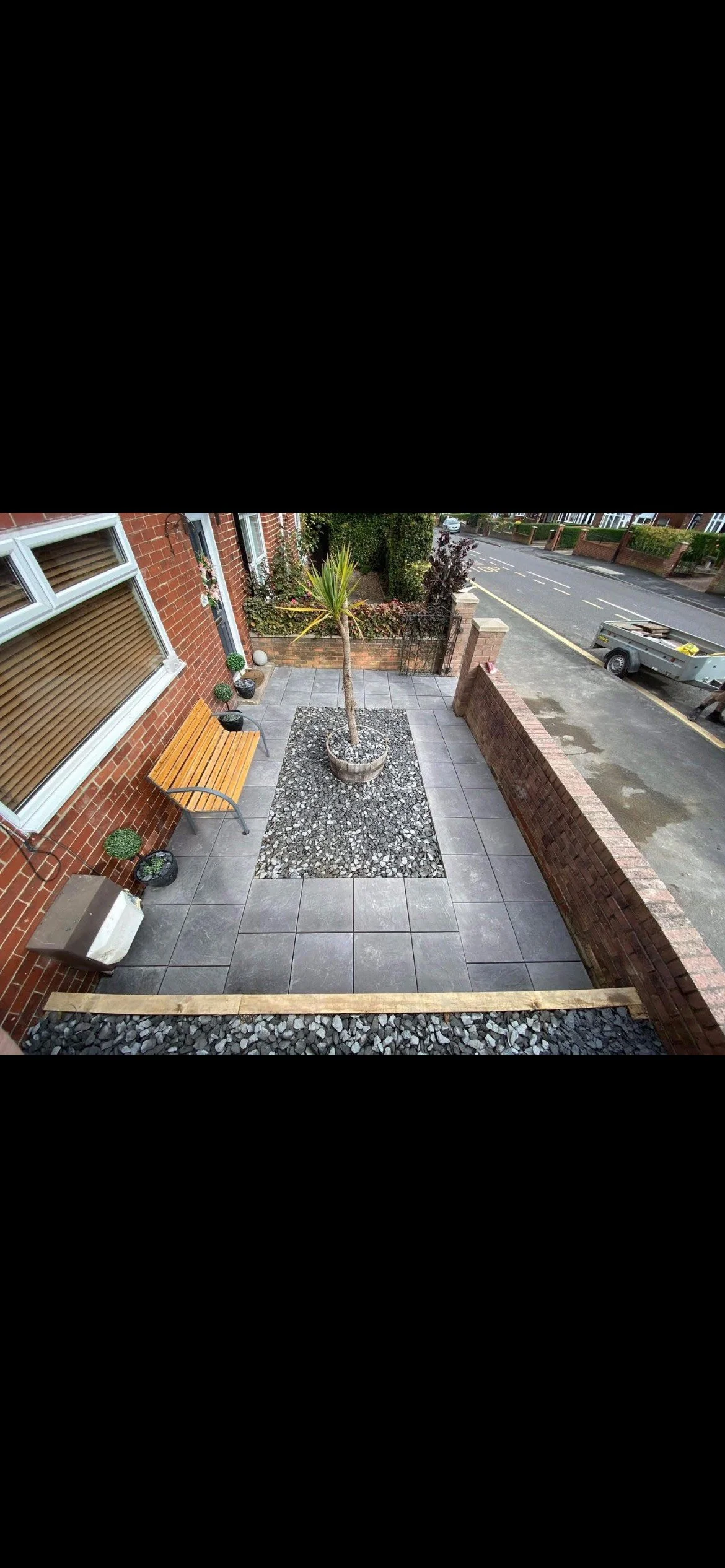 A front porch with large grey tiled flooring, a small tree in a pot, a wooden bench, potted plants, and a brick wall separating the porch from the street.