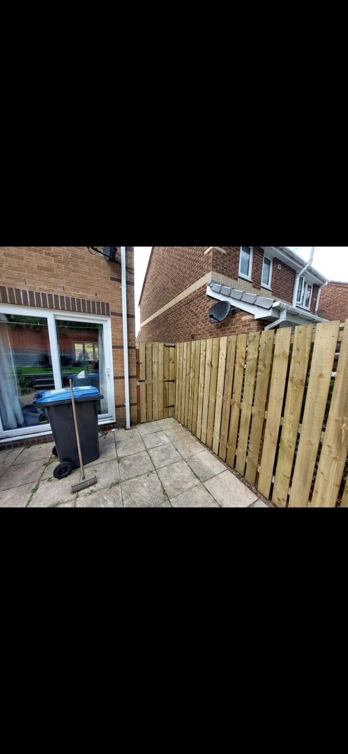 A small backyard patio with concrete tiles, a wheelie bin with a blue lid, a broom, and a wooden gate, enclosed by a wooden fence, with brick houses and a satellite dish in the background.