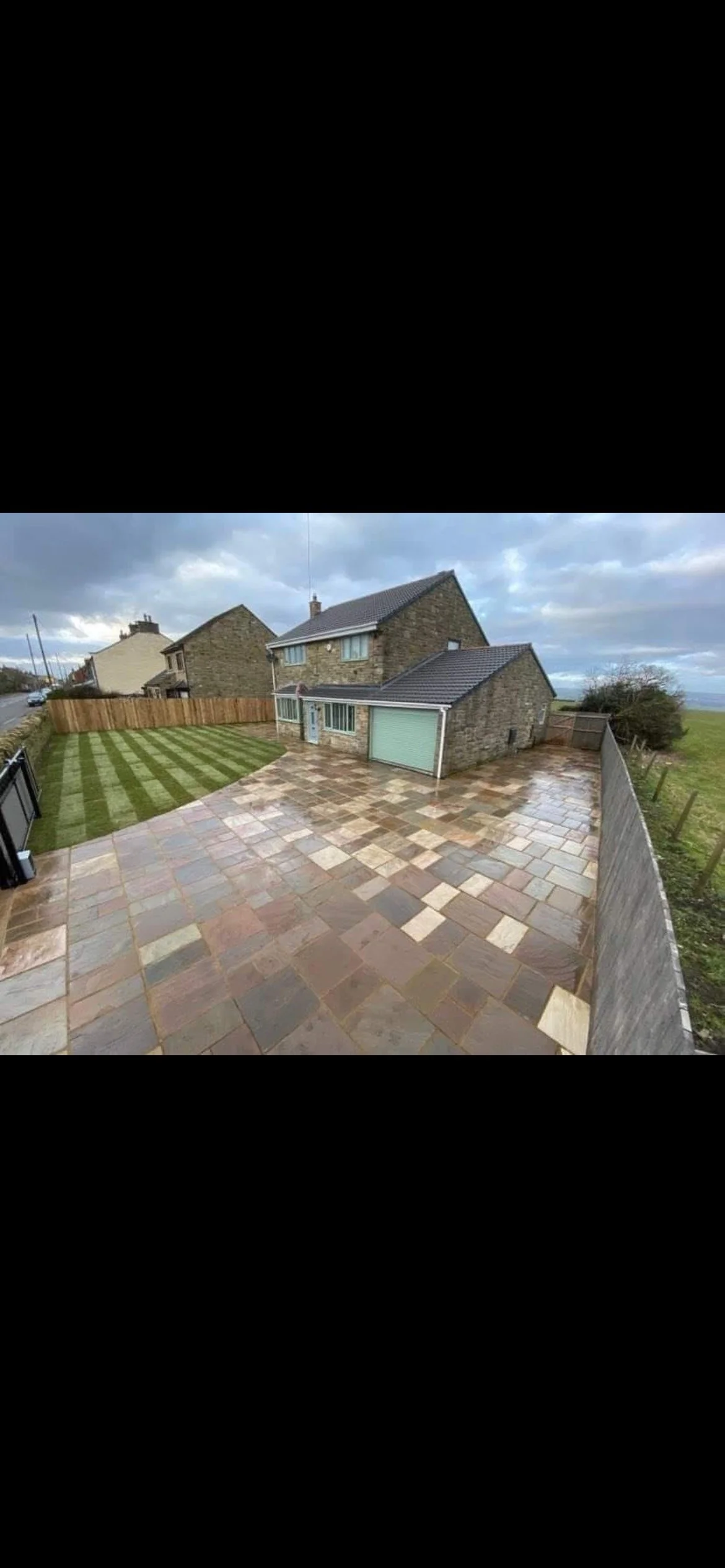 Photo of a backyard with a stone house, a paved patio, a grassy area with stripes, and a cloudy sky.