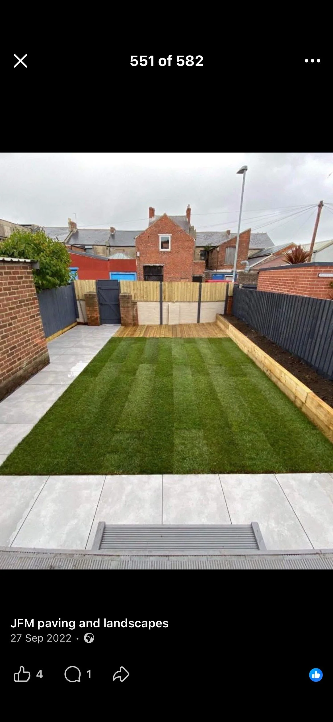 A backyard with a newly laid lawn, a concrete patio, and a wooden deck at the back. Brick and wood fencing surrounds the yard, with neighboring brick buildings in the background.