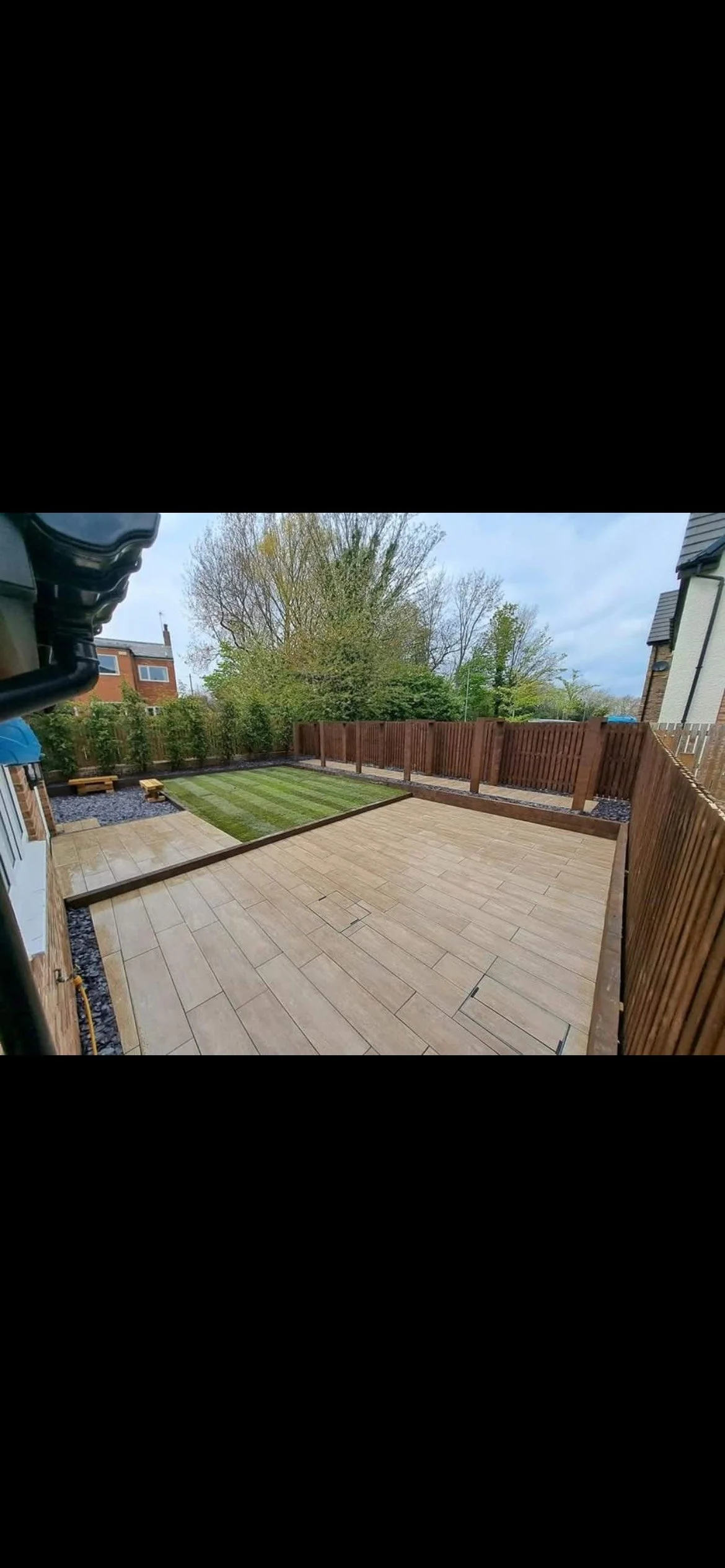 A newly landscaped backyard with a wooden fence, featuring a large paved patio area and a small grass lawn section, with trees in the background.