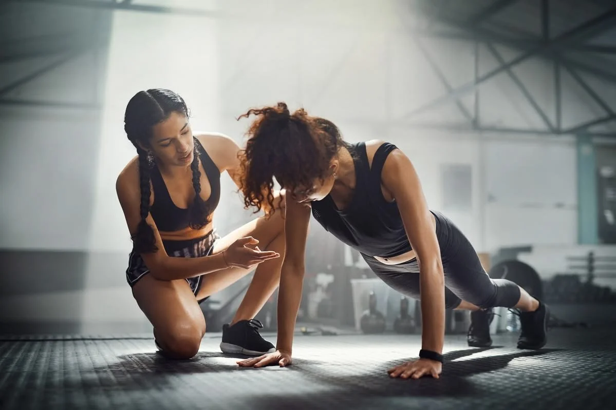 A woman doing a plank exercise in a gym with a fitness trainer providing guidance.