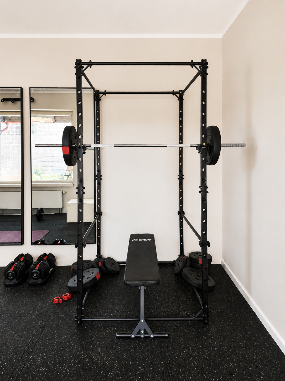 Home gym setup including a black power rack with an empty barbell and weight plates, a black workout bench, dumbbells, weight plates, and a large mirror on the wall.