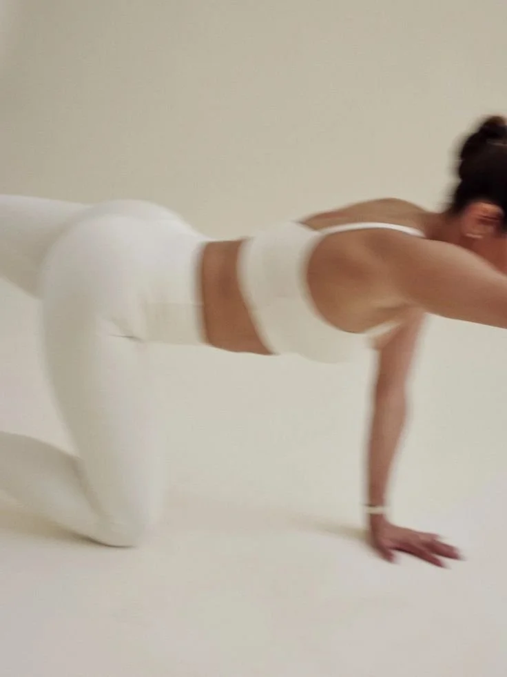 A person in white workout clothing is on all fours, performing an exercise on a flat, white surface in front of a plain, light-colored wall.