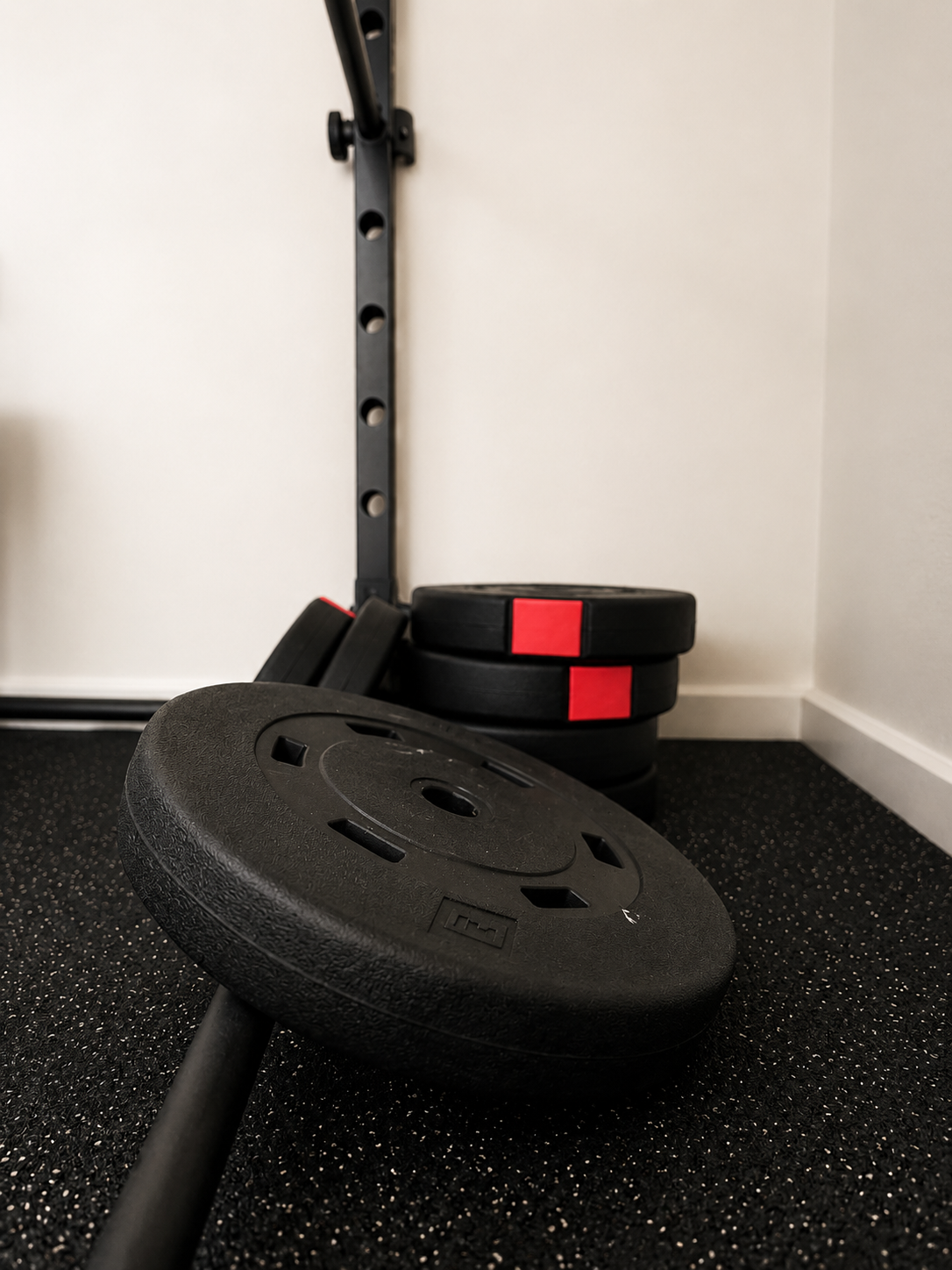 Black weight plate on the floor in front of a stack of similar weight plates with red stickers, with a black wall-mounted pull-up bar in the background.