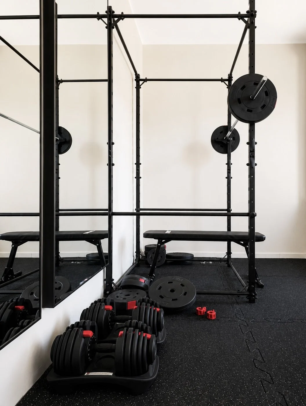 Empty gym corner with black weightlifting rack, bench, weight plates, dumbbells, and small red resistance bands.