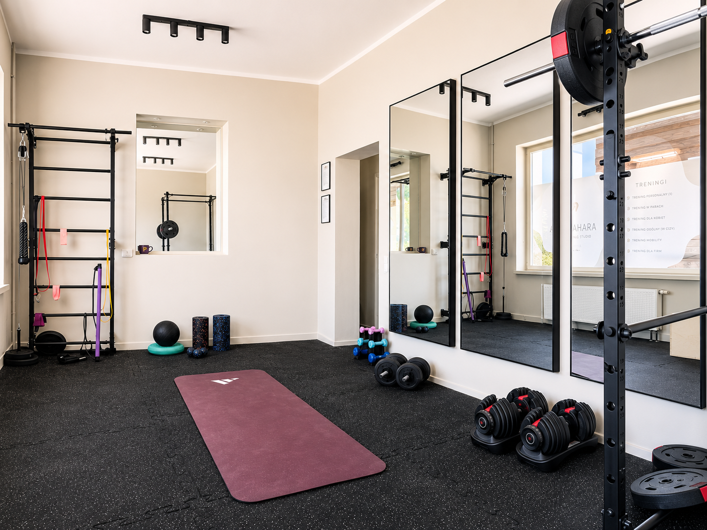 A home gym with various exercise equipment including a purple exercise mat, dumbbells, kettlebells, resistance bands, a stability ball, mirrors on the wall, and a wall-mounted pull-up bar with resistance bands attached. The room has black rubber floo