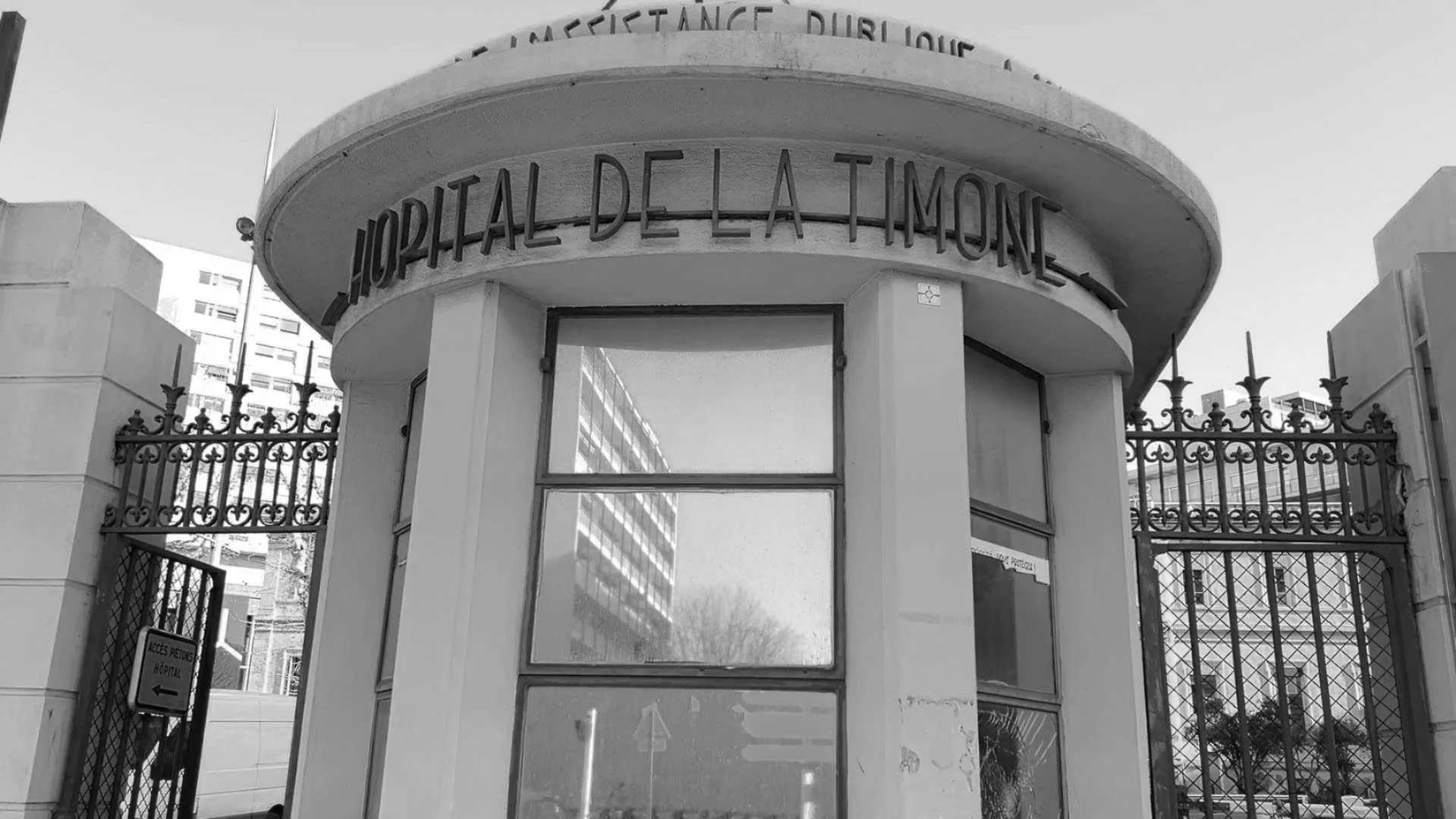 Entrée principale de l'hôpital de la Timone avec portail en fer forgé et un panneau d'indication. Vue en noir et blanc.
