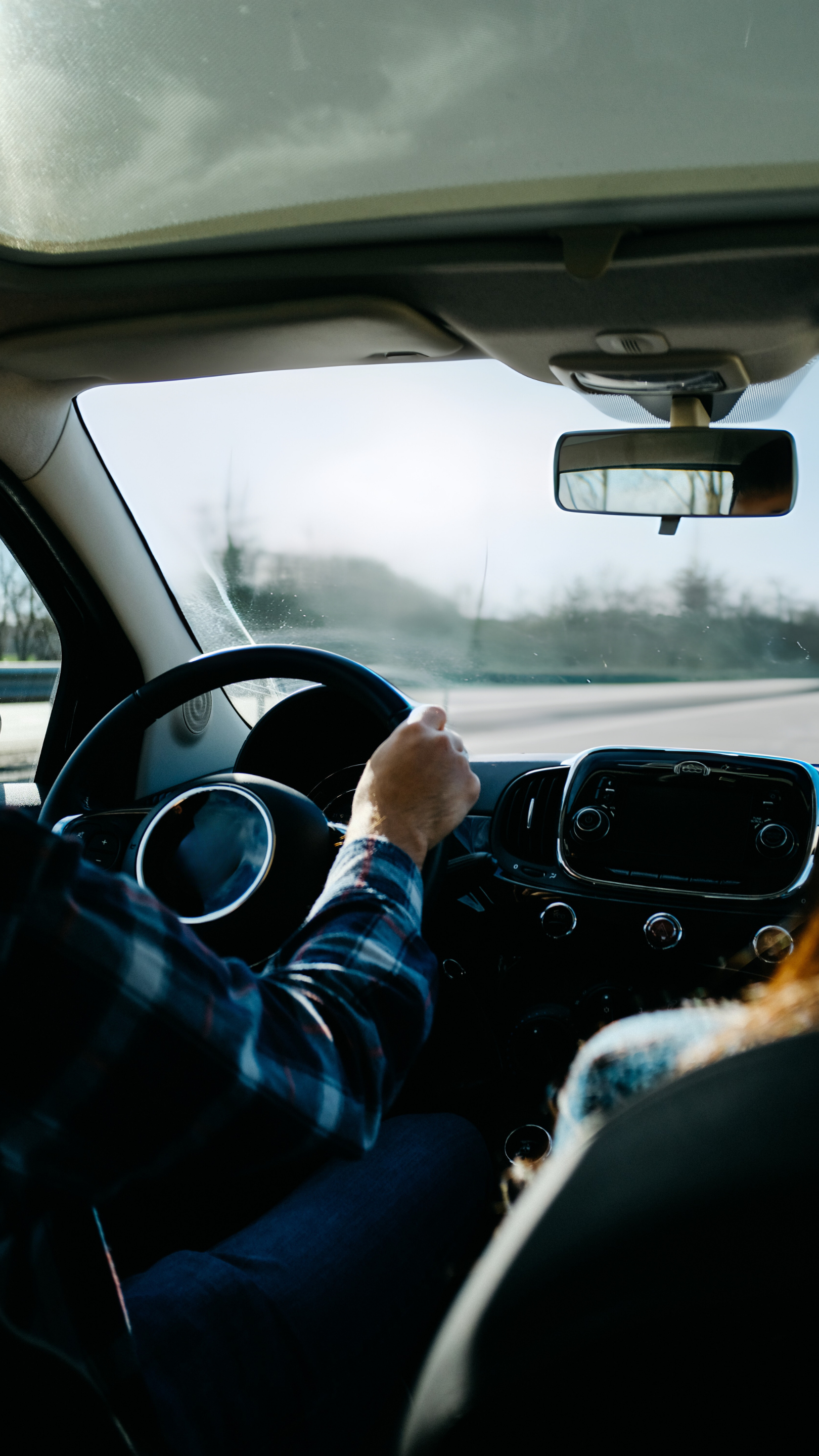 Vue de l'intérieur d'une voiture avec un conducteur portant une chemise à carreaux, vue de l'arrière, avec le tableau de bord et le miroir rétrovisé visible.