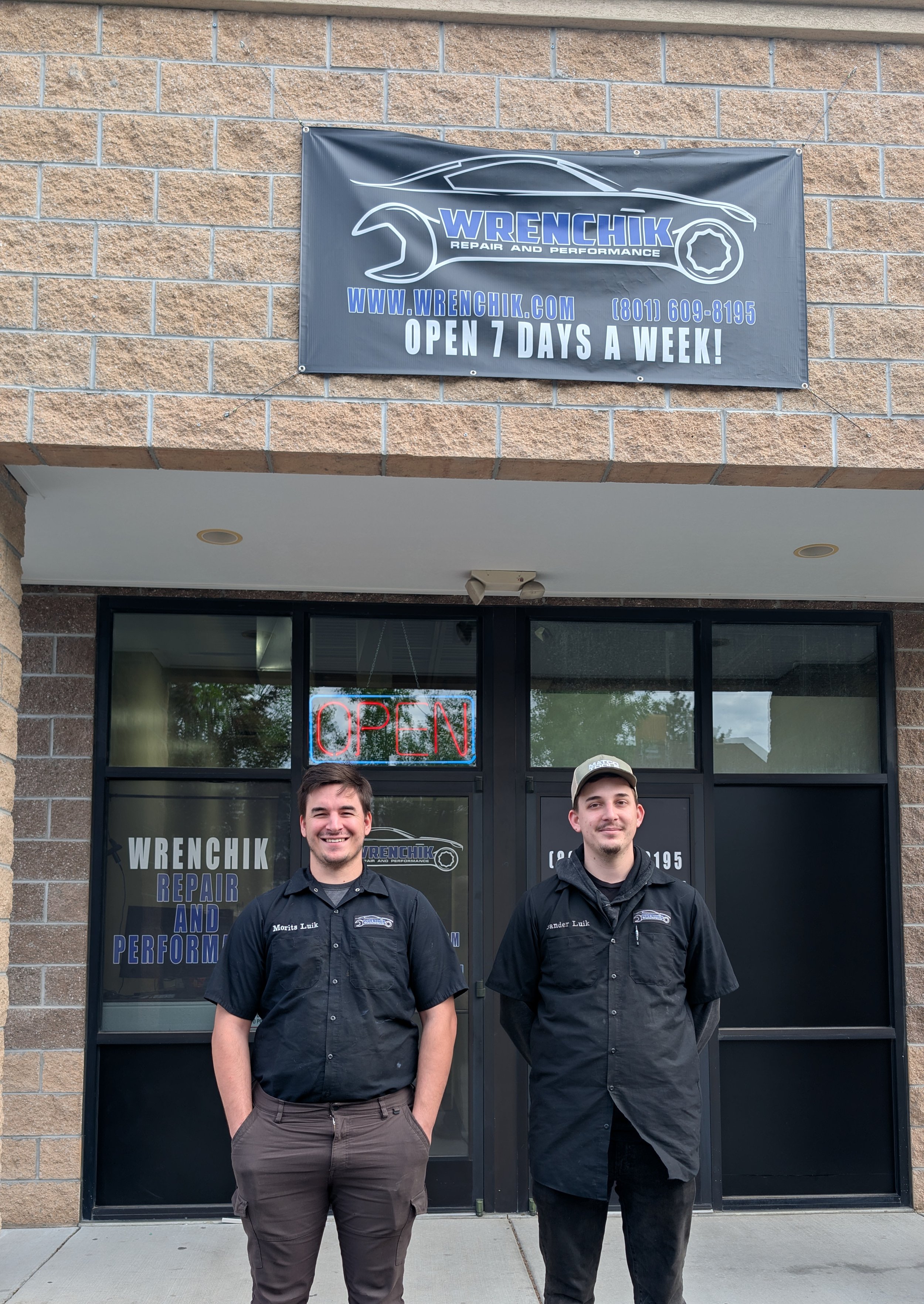 Two men standing in front of a business building with a sign for Wrenchik Repair and Performance. The man on the left has short dark hair, wearing a black shirt with the business logo, and has his hands in his pockets. The man on the right has short hair, wearing a black shirt with the business logo, and has his arms at his sides. Both are smiling.