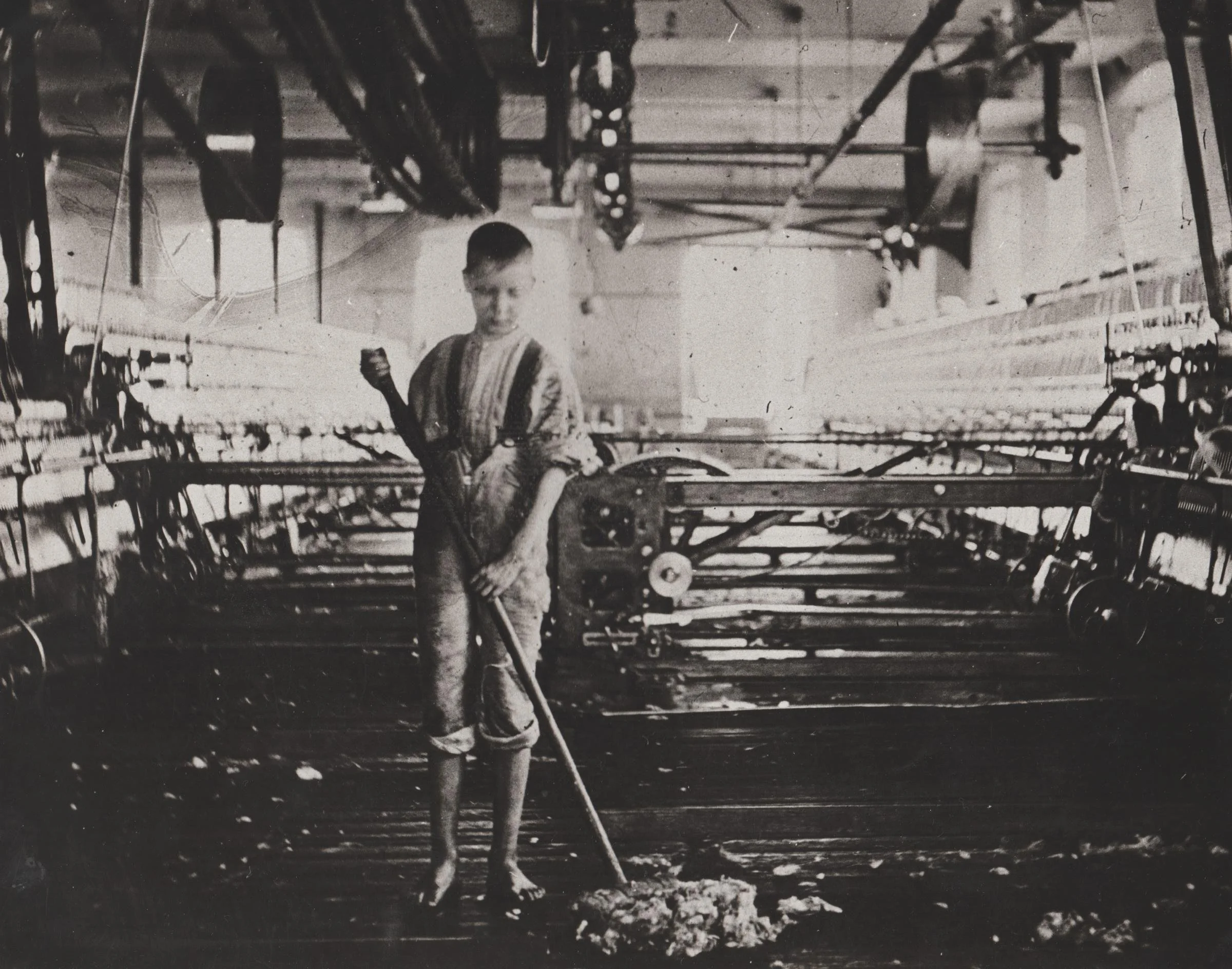 A young boy in old-fashioned clothing standing barefoot in a factory or workshop, holding a long rod or tool, surrounded by machinery and equipment.