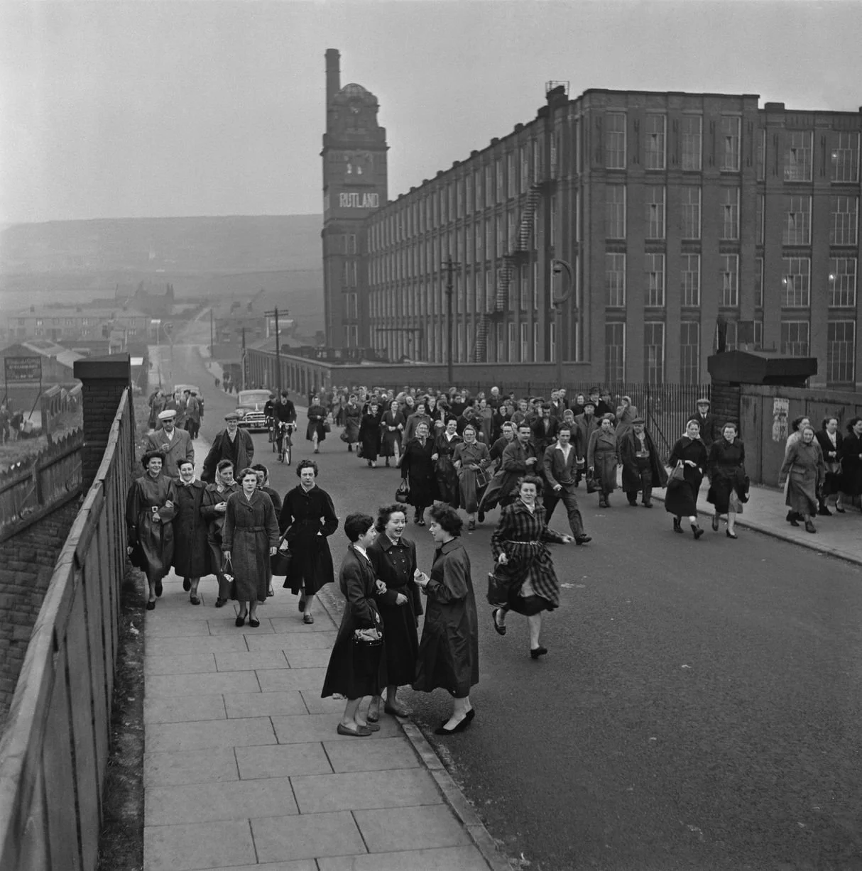 Black and white photo of a large group of women walking on a city street, with some Talking and others walking. The street is lined with tall brick buildings and a factory with a chimney is visible in the background.