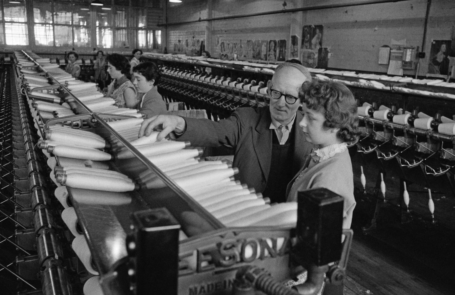 A man and a young girl observing textile machines in a factory, with other women working in the background.